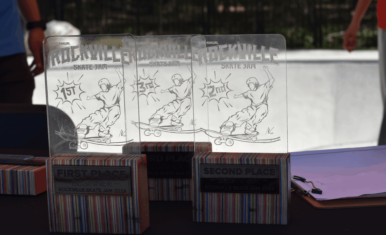 Three engraved glass trophies labeled First, Second, and Third Place for the Beginner division at the Rockville Skate Jam 2024 are displayed on a table outdoors.