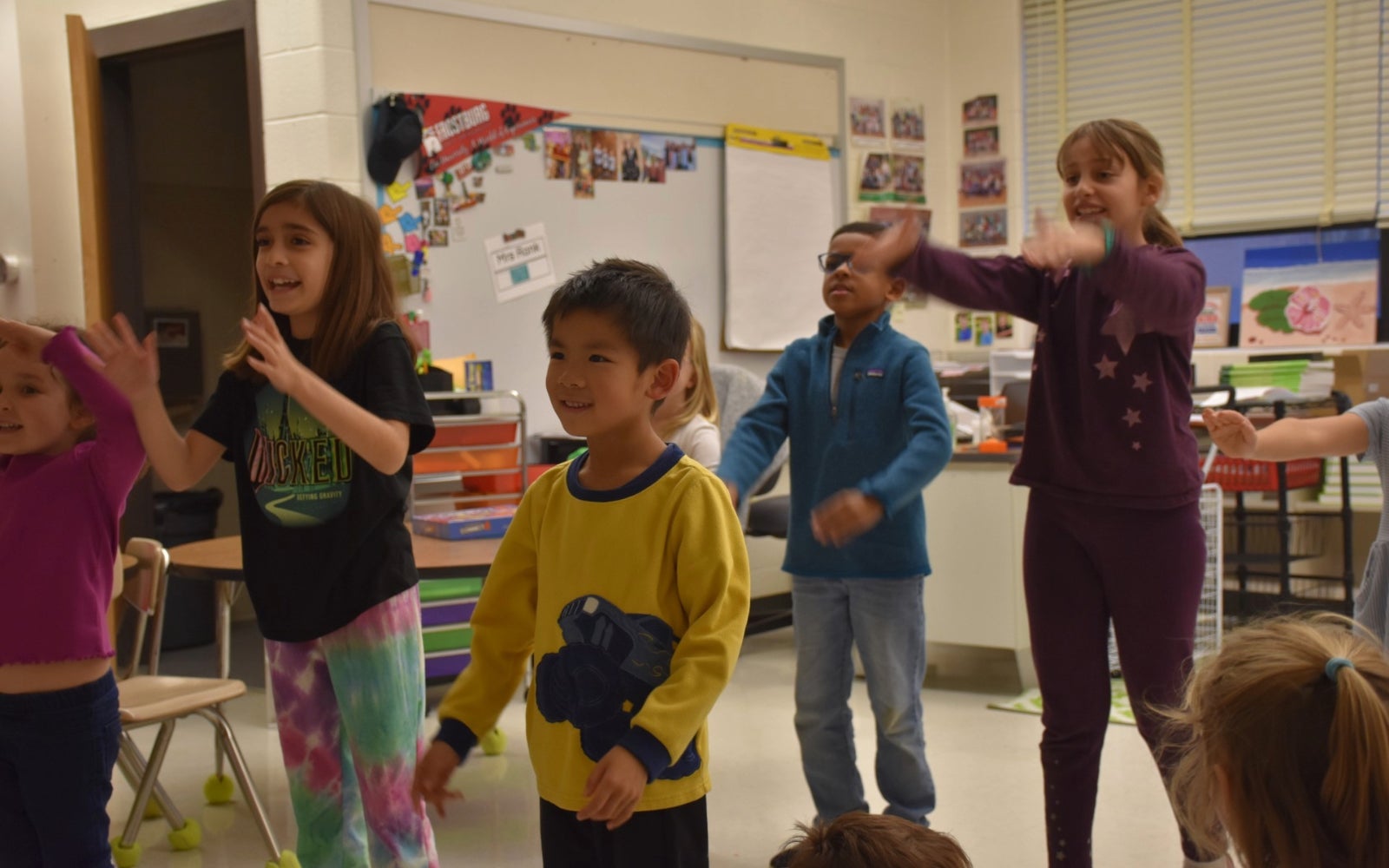 A group of young children stands in a classroom, smiling and raising their arms while participating in an activity together.