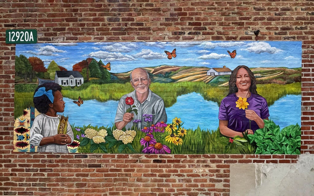 A mural on a brick wall shows a child, an older man, and a woman holding flowers in front of a lake, with butterflies, fields, and houses in the background.