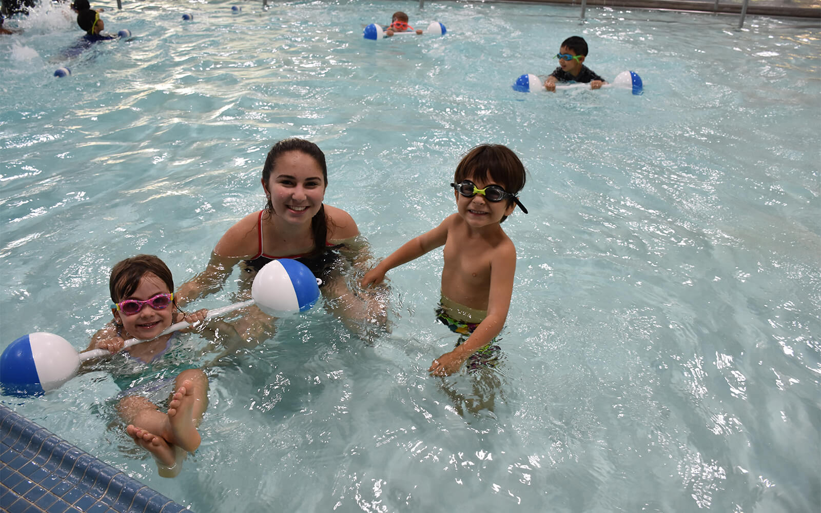 Three children and an adult are in a swimming pool, smiling at the camera. Two children hold blue and white floating balls, and all are wearing swim goggles.