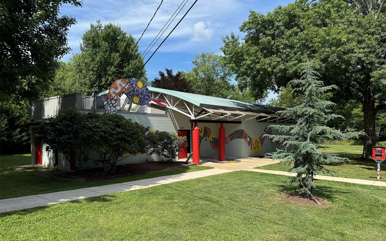 A small building with red columns and colorful mosaic art on the facade is surrounded by trees, grass, and a sidewalk under a blue sky.