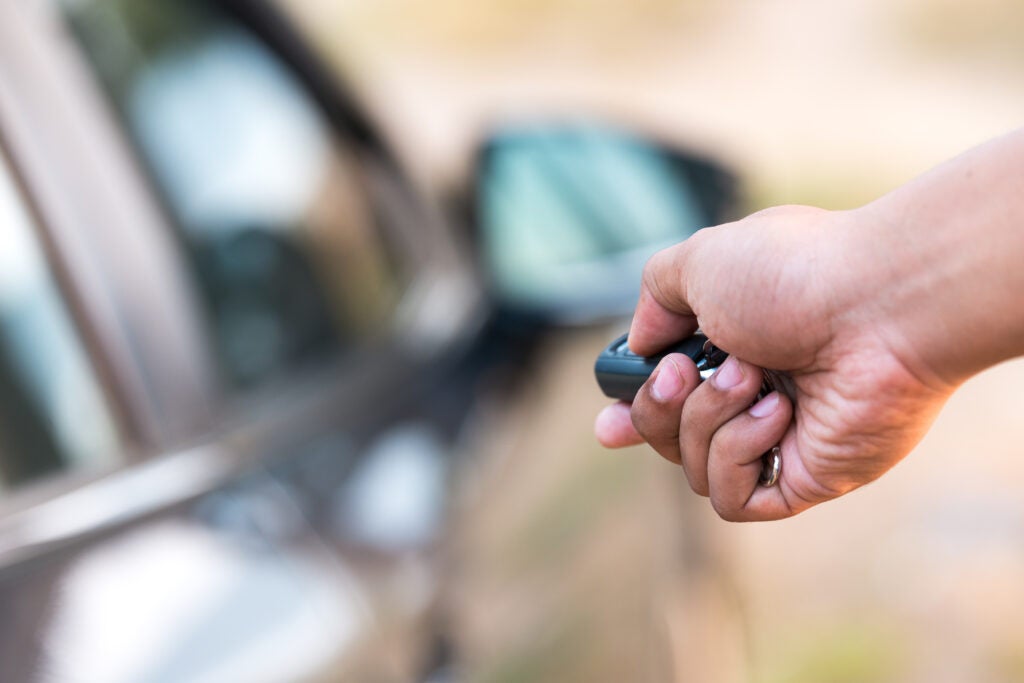 Close-up of a hand holding a car key fob, pointing it toward a parked car with a side mirror visible in the background.
