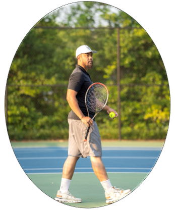 A man wearing a white cap, black shirt, and gray shorts holds a tennis racket and ball while walking on an outdoor tennis court.