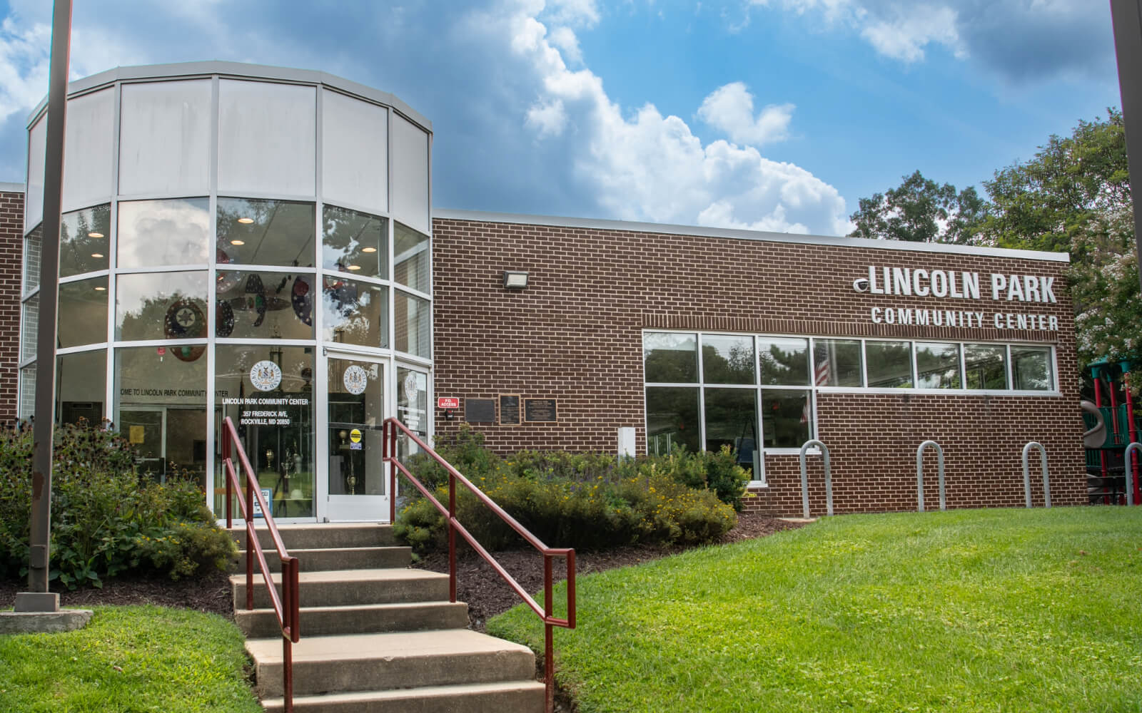 Exterior view of Lincoln Park Community Center with brick walls, glass entrance, concrete steps, handrails, and some landscaping on a partly cloudy day.