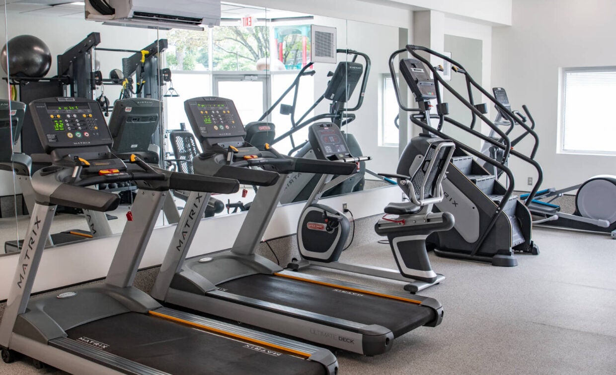 A gym with treadmills, an elliptical machine, and exercise bikes arranged in front of a wall mirror. Natural light enters through a window.