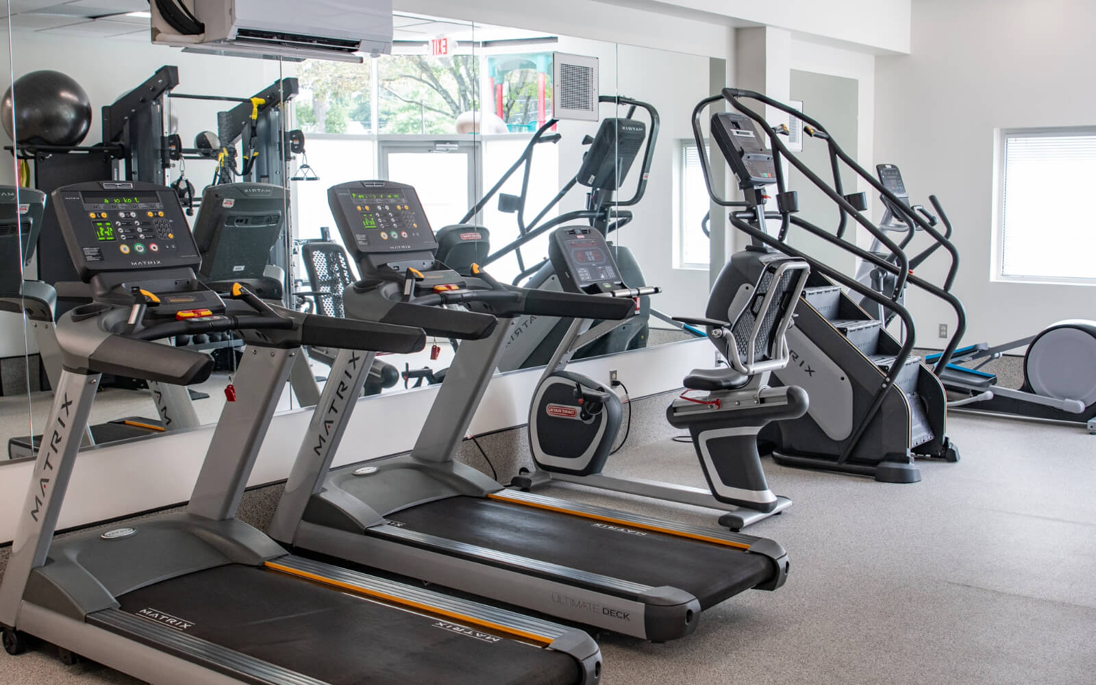 A gym with treadmills, an elliptical machine, and exercise bikes arranged in front of a wall mirror. Natural light enters through a window.