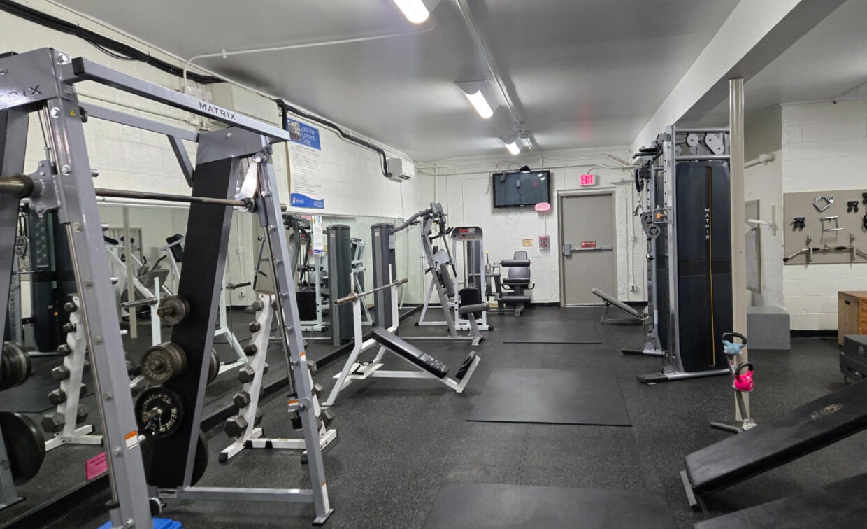 A gym with various exercise machines, weight racks, benches, and mats. The room is well-lit with white walls, black flooring, and a TV mounted on the wall.