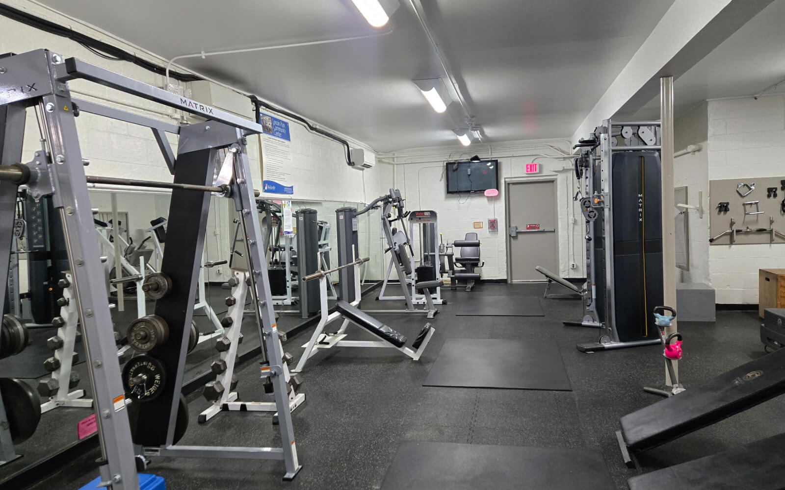A gym with various exercise machines, weight racks, benches, and mats. The room is well-lit with white walls, black flooring, and a TV mounted on the wall.