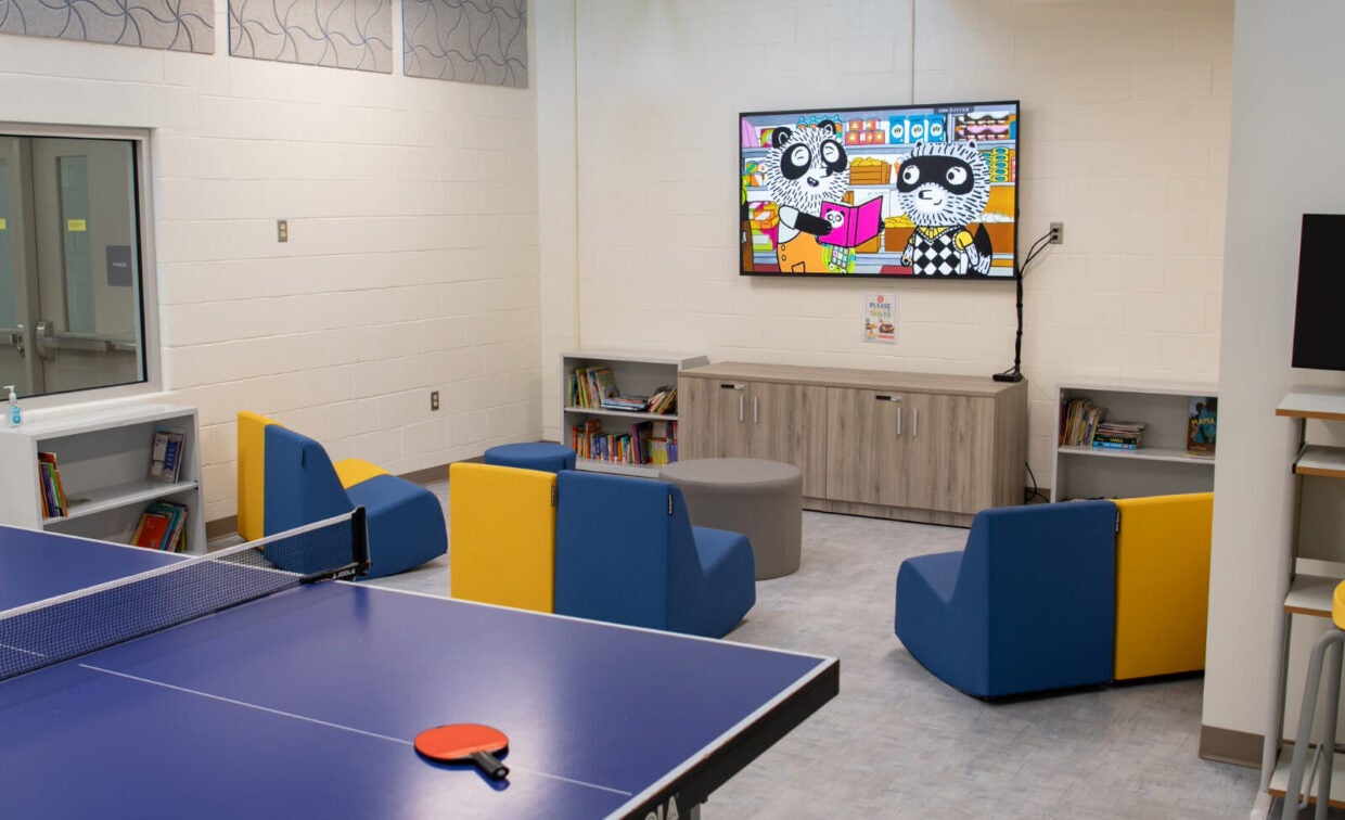 A modern children’s lounge with blue and yellow chairs, bookshelves, a TV displaying a cartoon, and a ping pong table in the foreground.