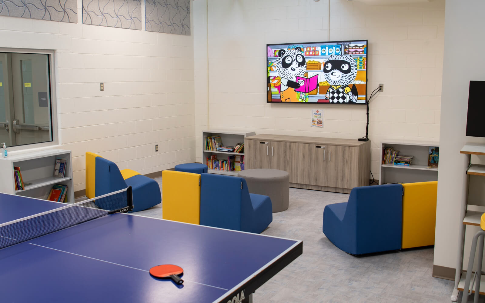 A modern children’s lounge with blue and yellow chairs, bookshelves, a TV displaying a cartoon, and a ping pong table in the foreground.