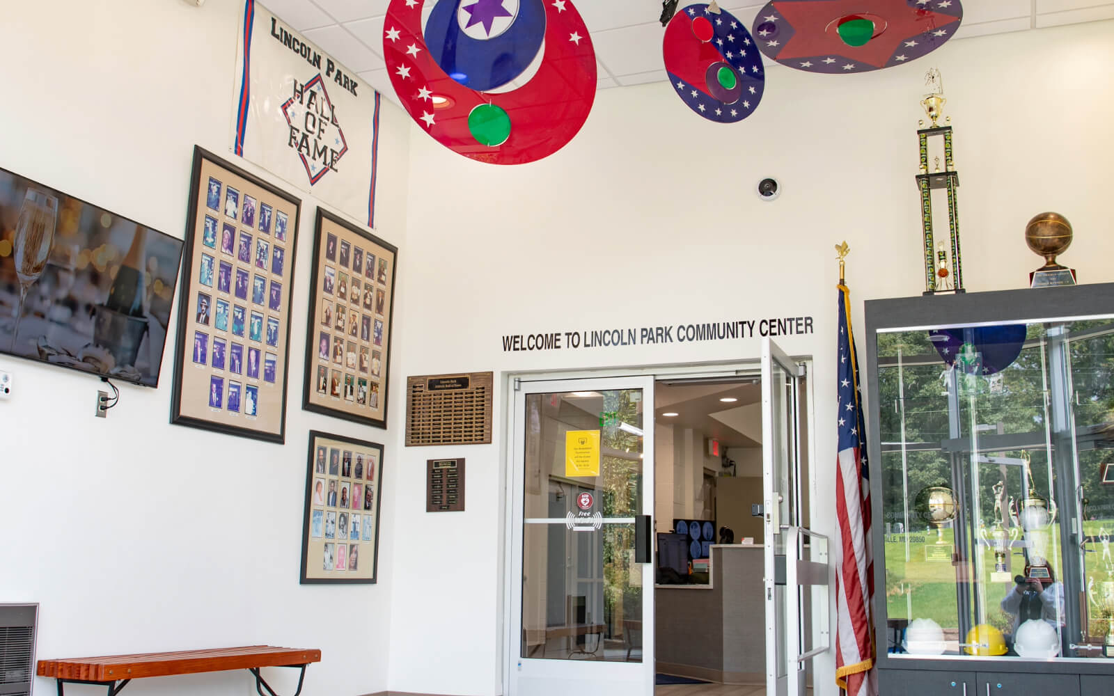 Lobby of a community center with trophy case, framed photos, plaques, American flag, and decorative ceiling art. Sign above doors reads “Welcome to Lincoln Park Community Center.”.