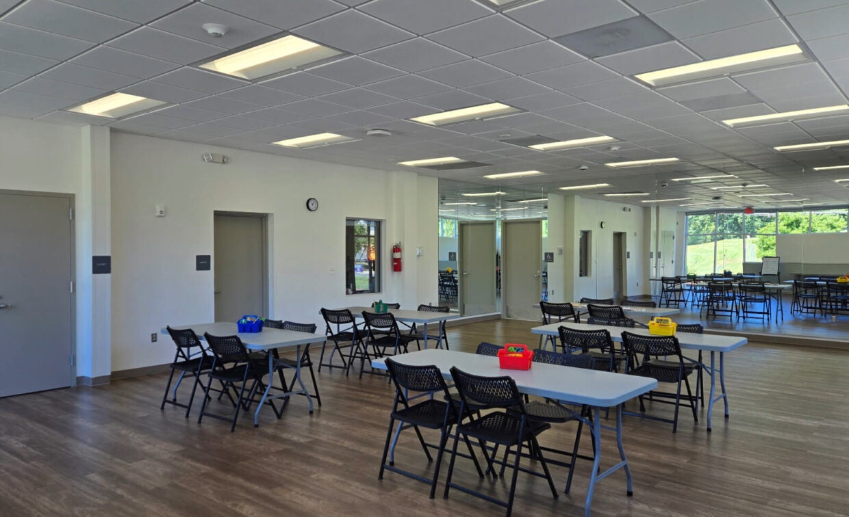 A bright, modern classroom with folding tables and chairs, tile ceiling, wood floor, large mirrors, and windows letting in natural light.