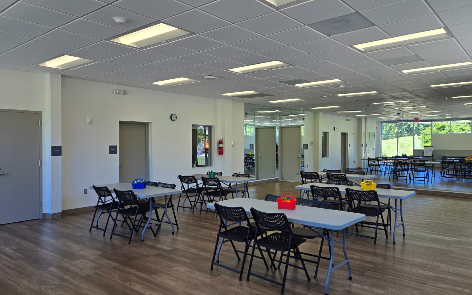 A bright, modern classroom with folding tables and chairs, tile ceiling, wood floor, large mirrors, and windows letting in natural light.