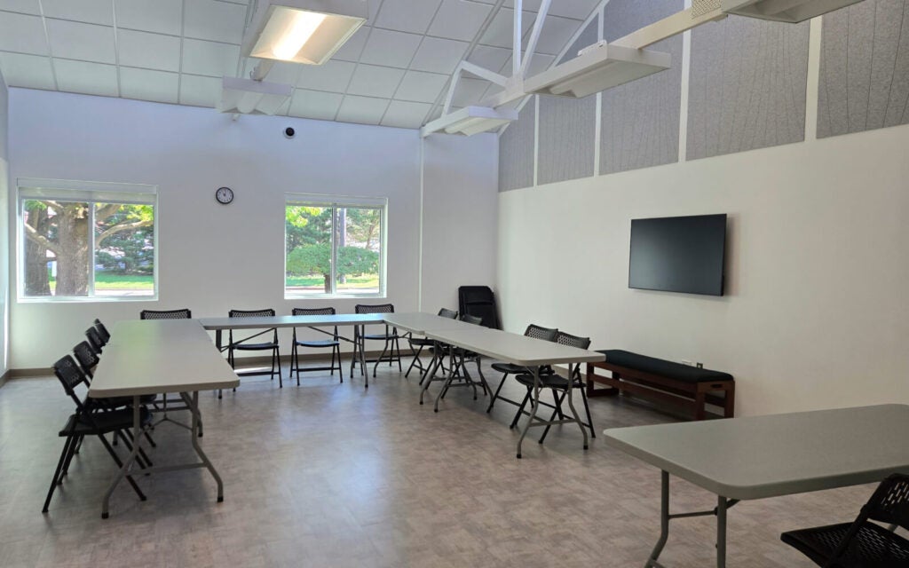 A conference room with folding tables and chairs arranged in a U-shape, a wall-mounted TV, clock, and windows showing trees outside.