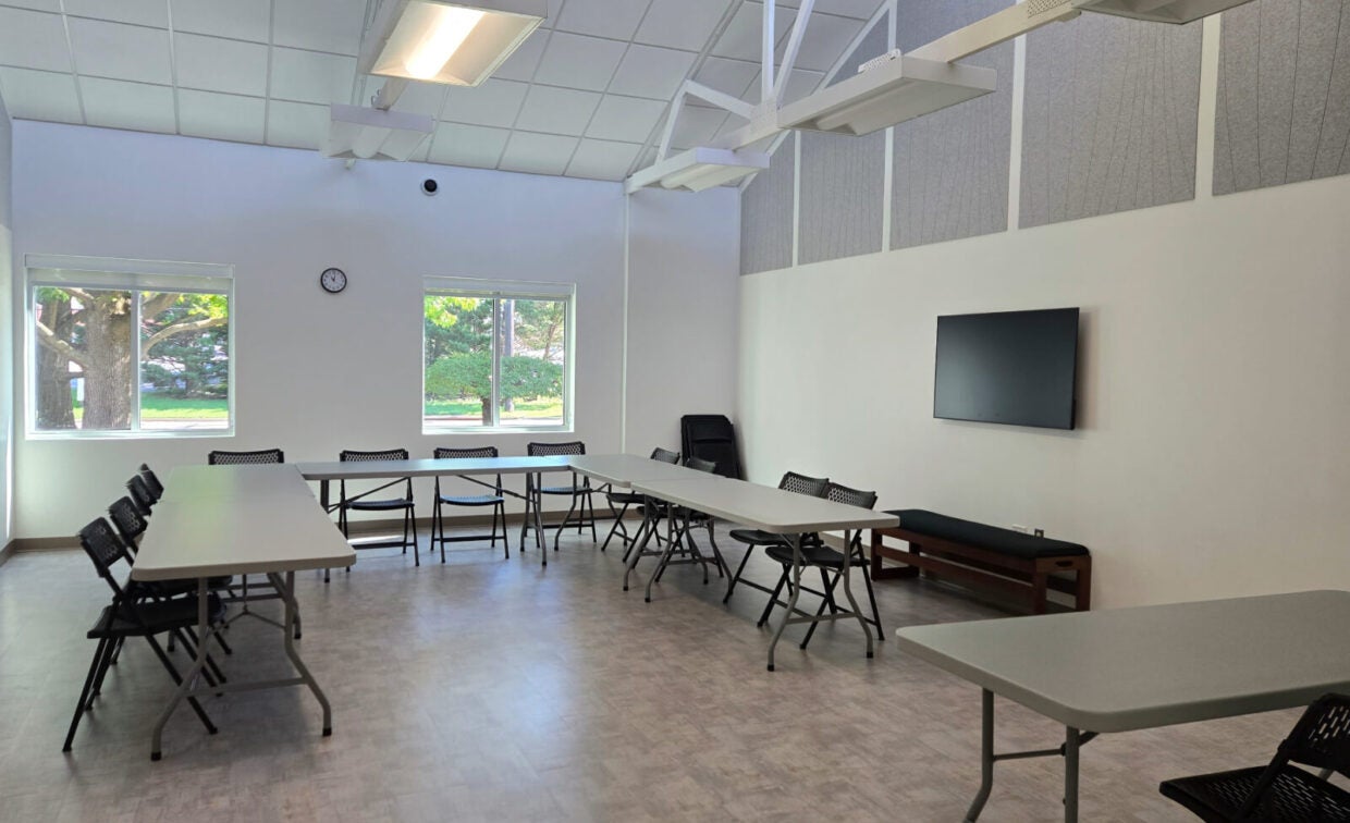 A conference room with folding tables and chairs arranged in a U-shape, a wall-mounted TV, clock, and windows showing trees outside.