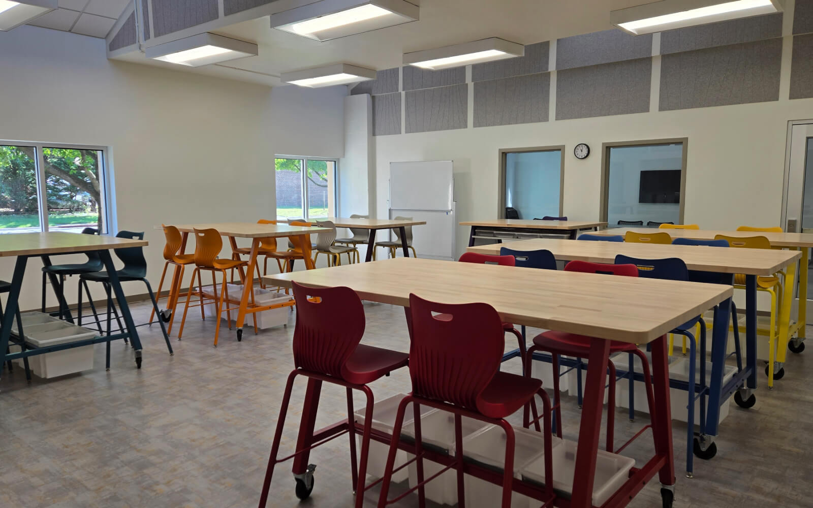 Classroom with high tables and colorful chairs arranged in groups, large windows, a whiteboard, and a clock on the wall. The room is well-lit with overhead lighting.