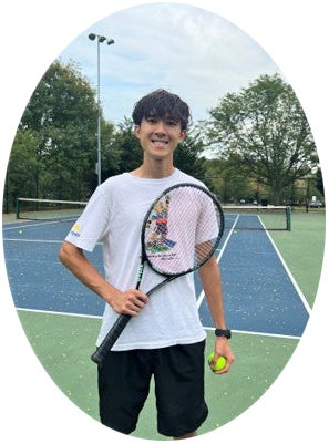 Young person standing on a tennis court, holding a racket and tennis ball, wearing a white shirt and black shorts, with trees and net visible in the background.