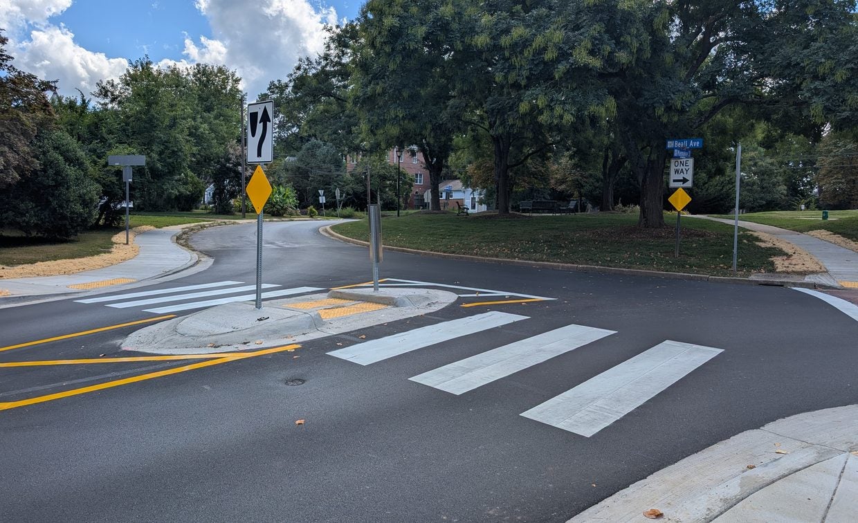 A roundabout intersection with a pedestrian crosswalk, traffic signs, and surrounding trees on a sunny day.