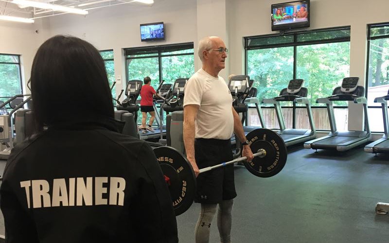 An older man lifts a barbell in a gym while a trainer observes. Treadmills and other people exercising are visible in the background.