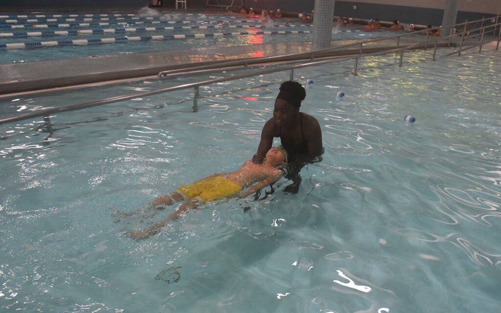 An adult helps a child in yellow shorts float on their back in a swimming pool during a swim lesson.