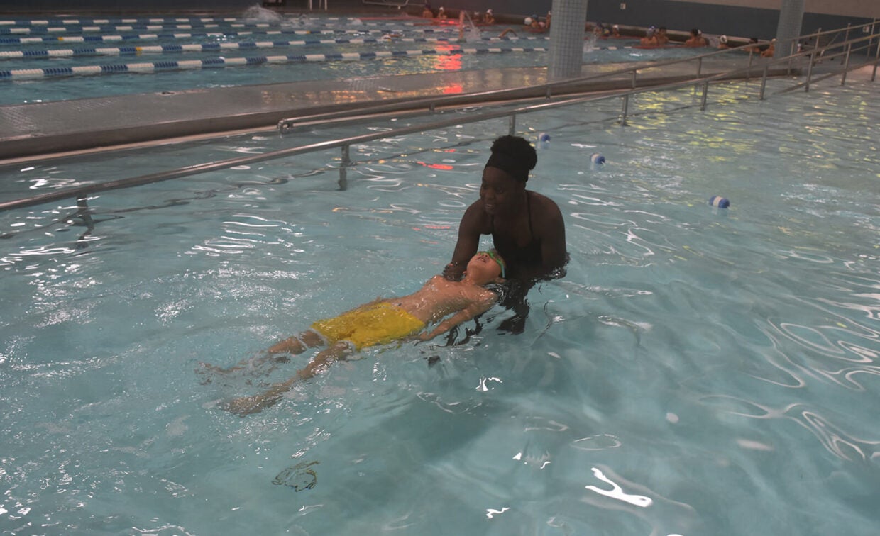 An adult helps a child in yellow shorts float on their back in a swimming pool during a swim lesson.