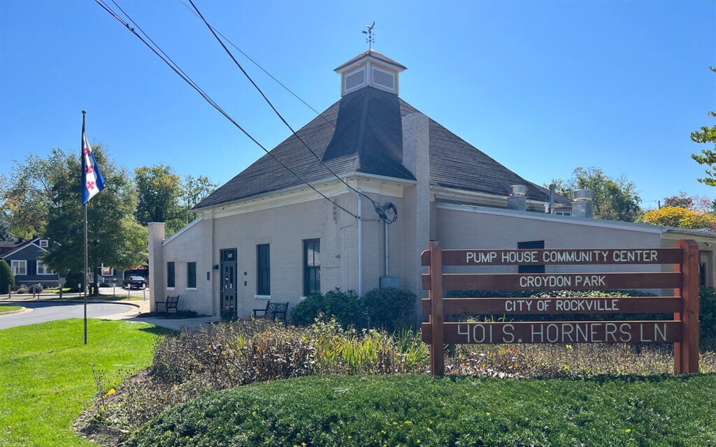 A beige building with a sign reading "Pump House Community Center, Croydon Park, City of Rockville, 401 S. Horners Ln" stands next to a flagpole and greenery on a sunny day.