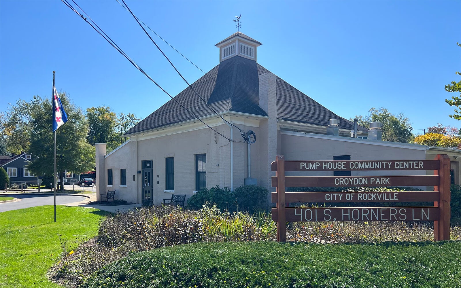 A beige building with a sign reading "Pump House Community Center, Croydon Park, City of Rockville, 401 S. Horners Ln" stands next to a flagpole and greenery on a sunny day.