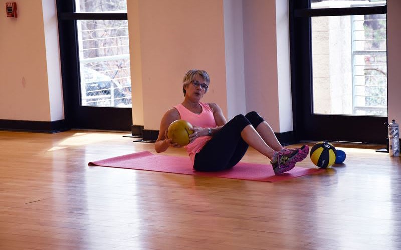 A woman sits on a yoga mat in a gym, holding a medicine ball and twisting her torso, with additional medicine balls nearby.