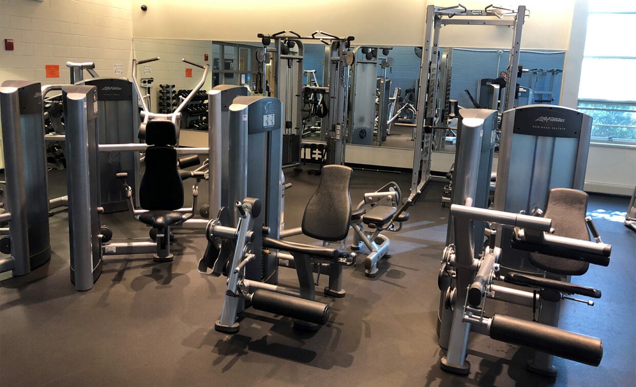 Several strength training machines arranged in a well-lit gym with mirrors and windows, including leg and chest press equipment on a clean floor.