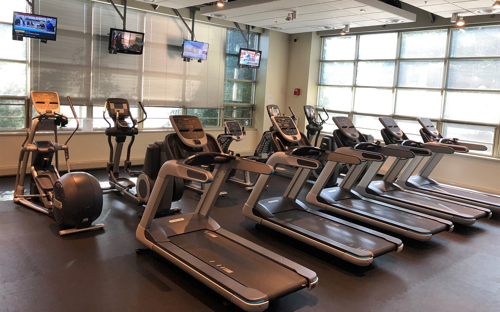 A gym with several treadmills and elliptical machines facing windows and wall-mounted TVs, in a well-lit, empty room.