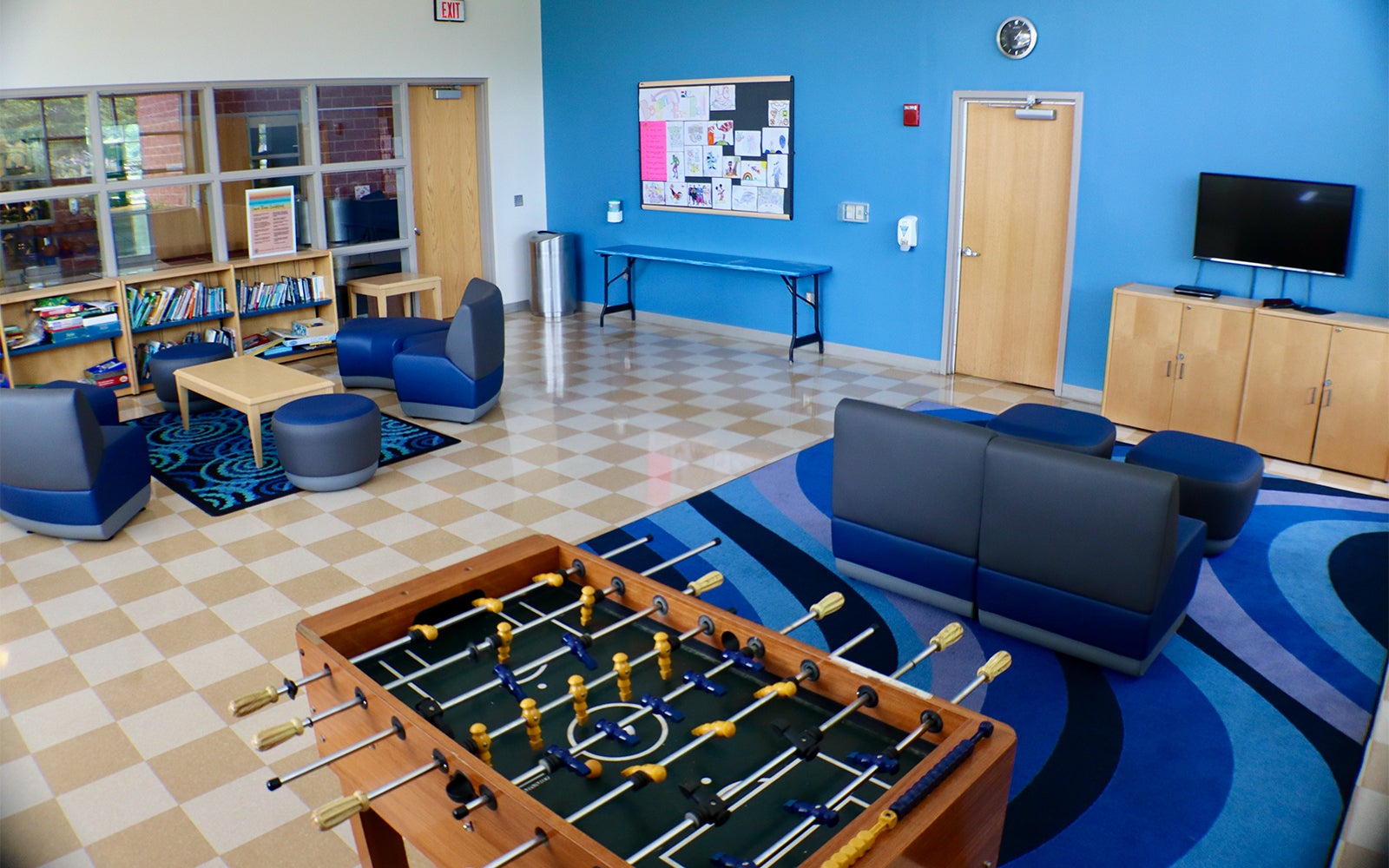 A recreation room with a foosball table, modern seating, bookshelves, a TV, a bulletin board, and large windows along one wall.