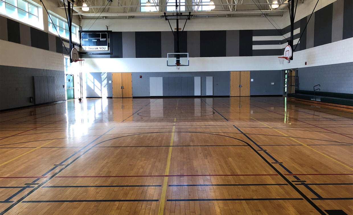 Empty indoor gymnasium with polished wooden floor, basketball hoops, scoreboard, and natural light streaming through windows on the left wall.