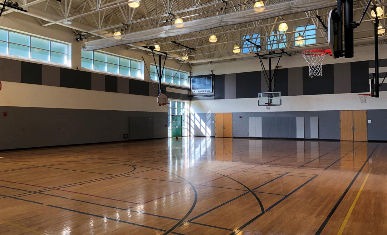 Empty indoor basketball gym with polished wooden floor, multiple hoops, high ceilings, and natural light coming through upper windows.