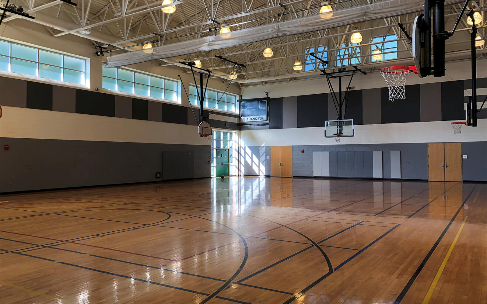 Empty indoor basketball gym with polished wooden floor, multiple hoops, high ceilings, and natural light coming through upper windows.