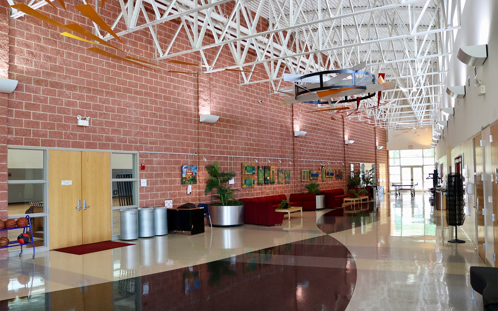 Modern school hallway with brick walls, colorful floor, plants, red couches, artwork on display, and suspended model airplanes from the ceiling.