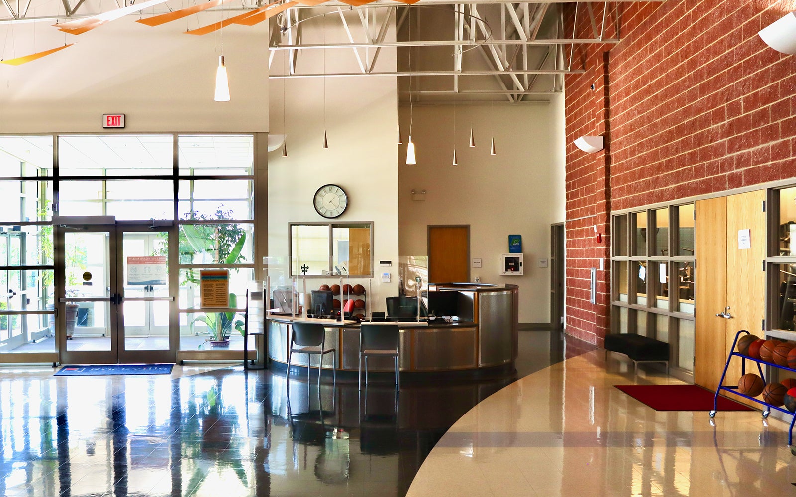 Spacious building lobby with glass entrance doors, a circular reception desk, wall clock, red brick wall, and windows to adjacent offices. Ceiling beams and orange ribbons hang overhead.