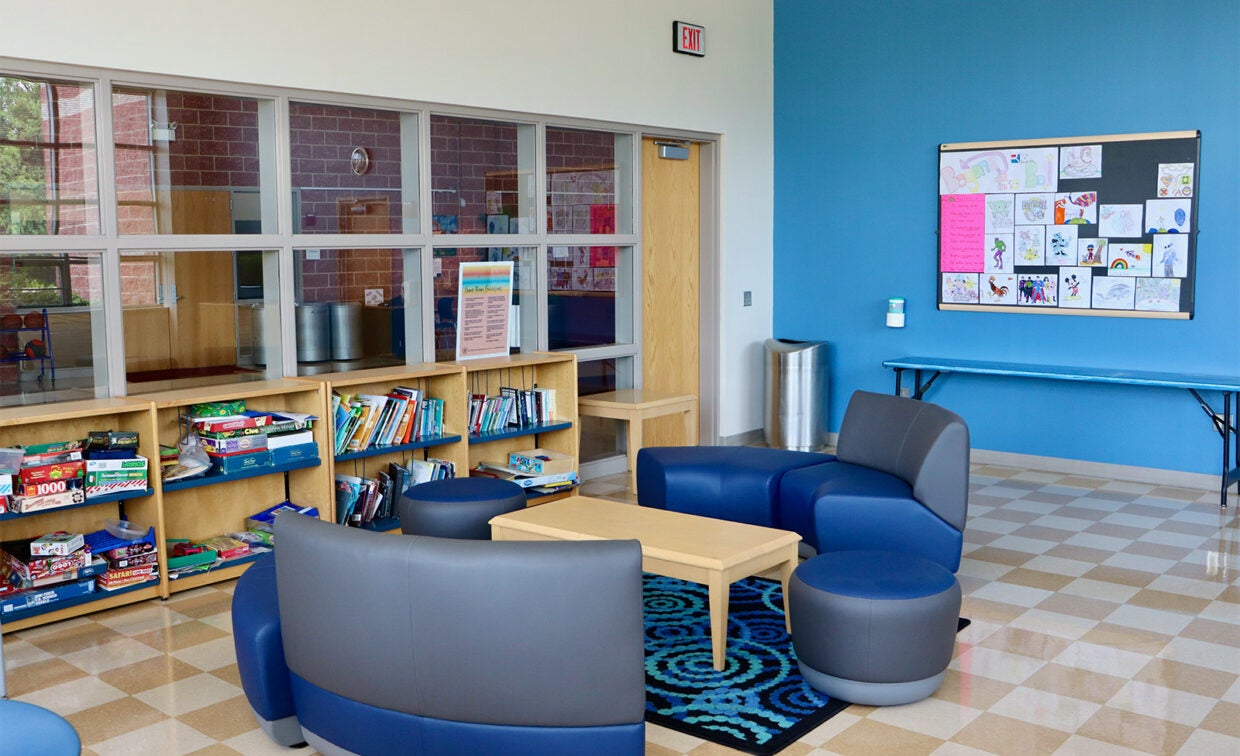A modern room with blue walls, bookshelves, board games, and round chairs around a small table. Children's drawings are displayed on a bulletin board.