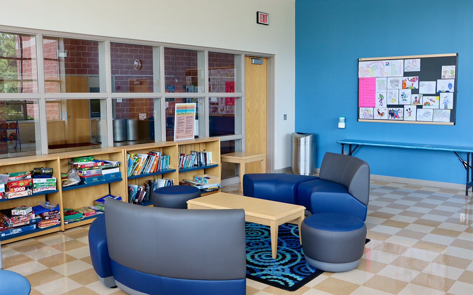 A modern room with blue walls, bookshelves, board games, and round chairs around a small table. Children's drawings are displayed on a bulletin board.