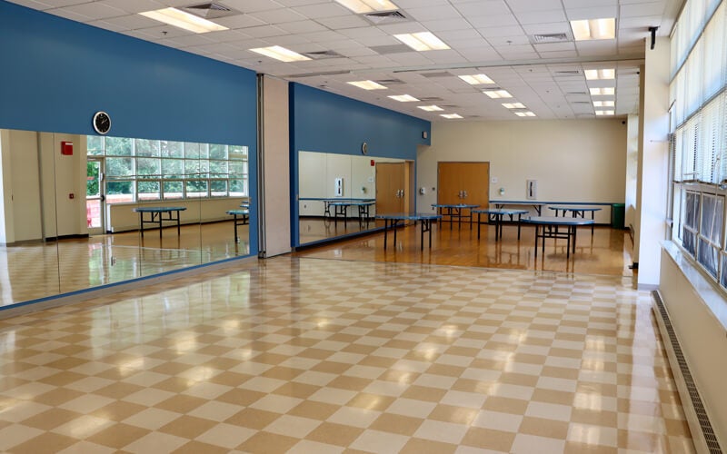 Large empty room with checkered floor, blue accent wall, mirrors, windows on one side, tables at the back, and fluorescent ceiling lights.
