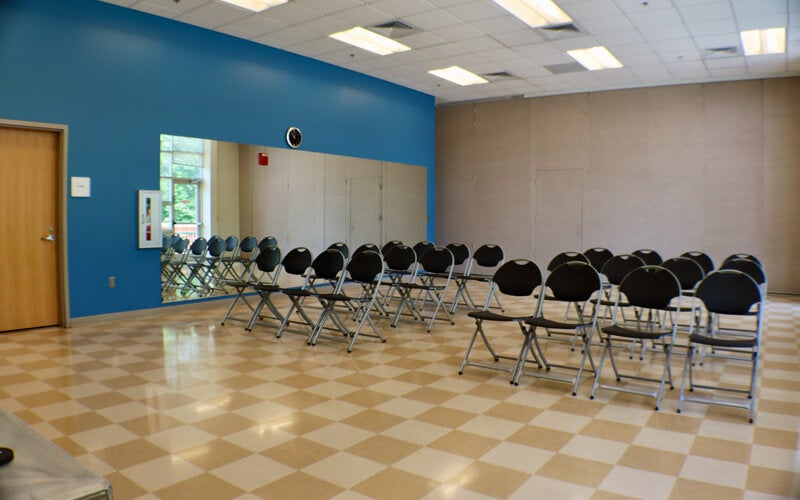 A room with a blue accent wall, large mirror, rows of black folding chairs facing forward, and a checkered tile floor under bright ceiling lights.