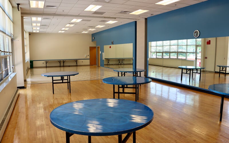 Large room with wood floors, blue walls, mirrors, and round blue tables arranged throughout. Bright lighting and large windows provide natural light.
