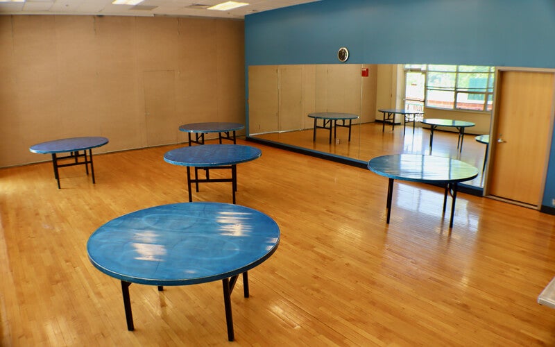 A room with five round blue tables on a wooden floor, blue and tan walls, a large wall mirror, a clock, and a window letting in natural light.