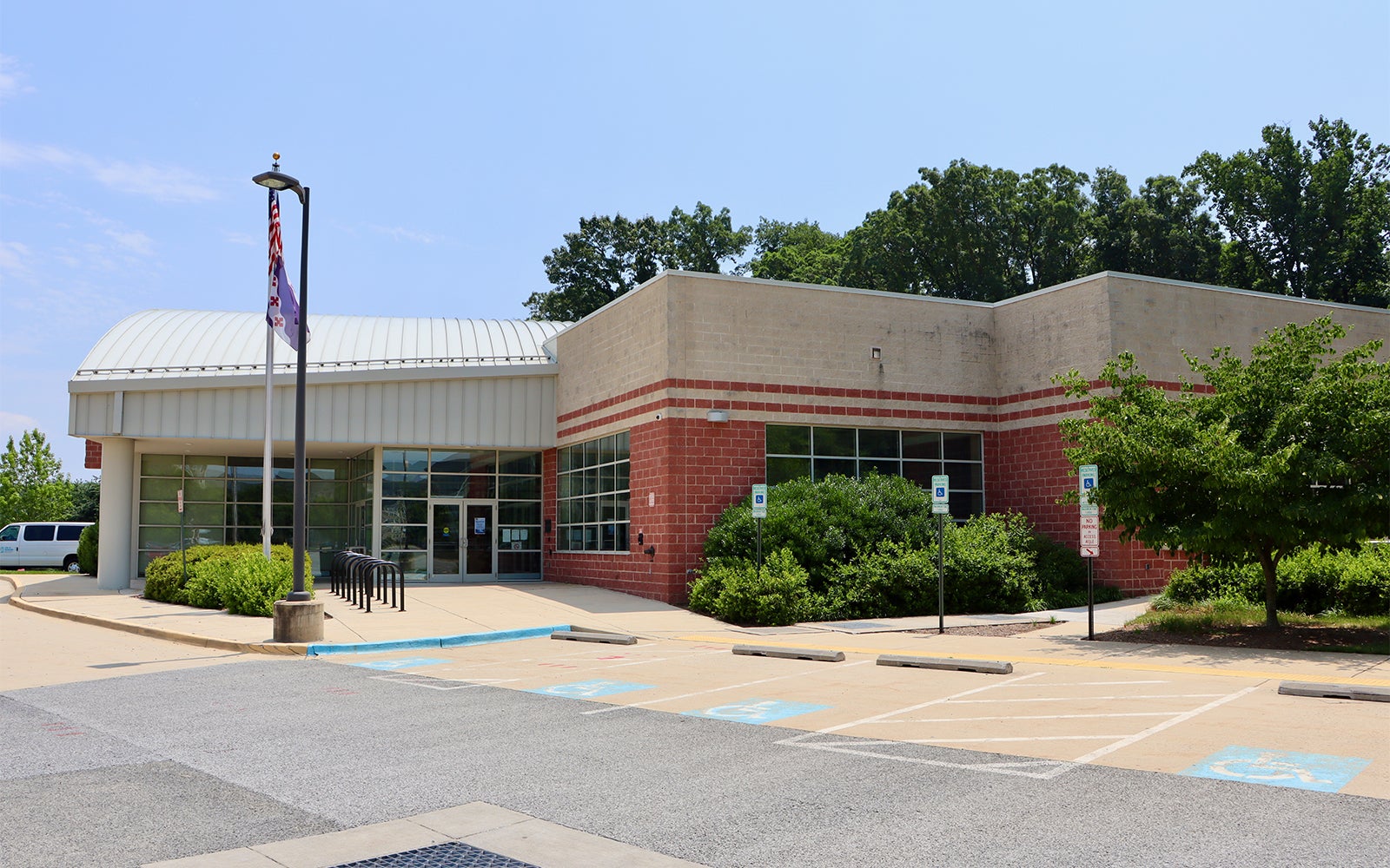 Single-story public building with brick and beige walls, large windows, glass entrance doors, and adjacent accessible parking spaces, surrounded by greenery.
