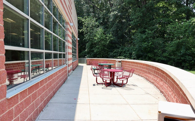 Outdoor patio with red tables and chairs next to a brick building, bordered by a curved wall and trees in the background.