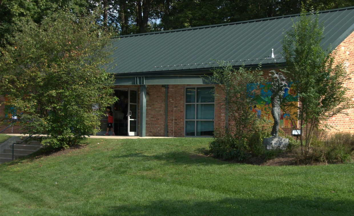 A brick building with a green metal roof, surrounded by grass and trees, has a mural on one wall and an open door with a person visible inside.
