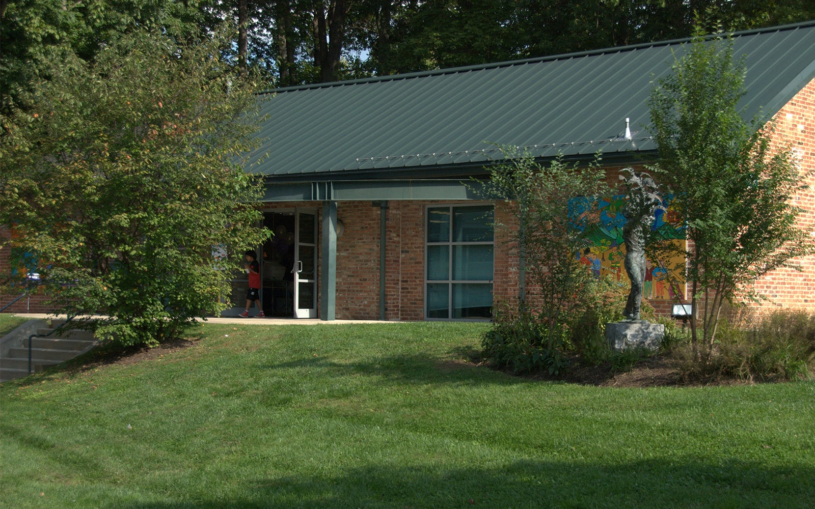A brick building with a green metal roof, surrounded by grass and trees, has a mural on one wall and an open door with a person visible inside.