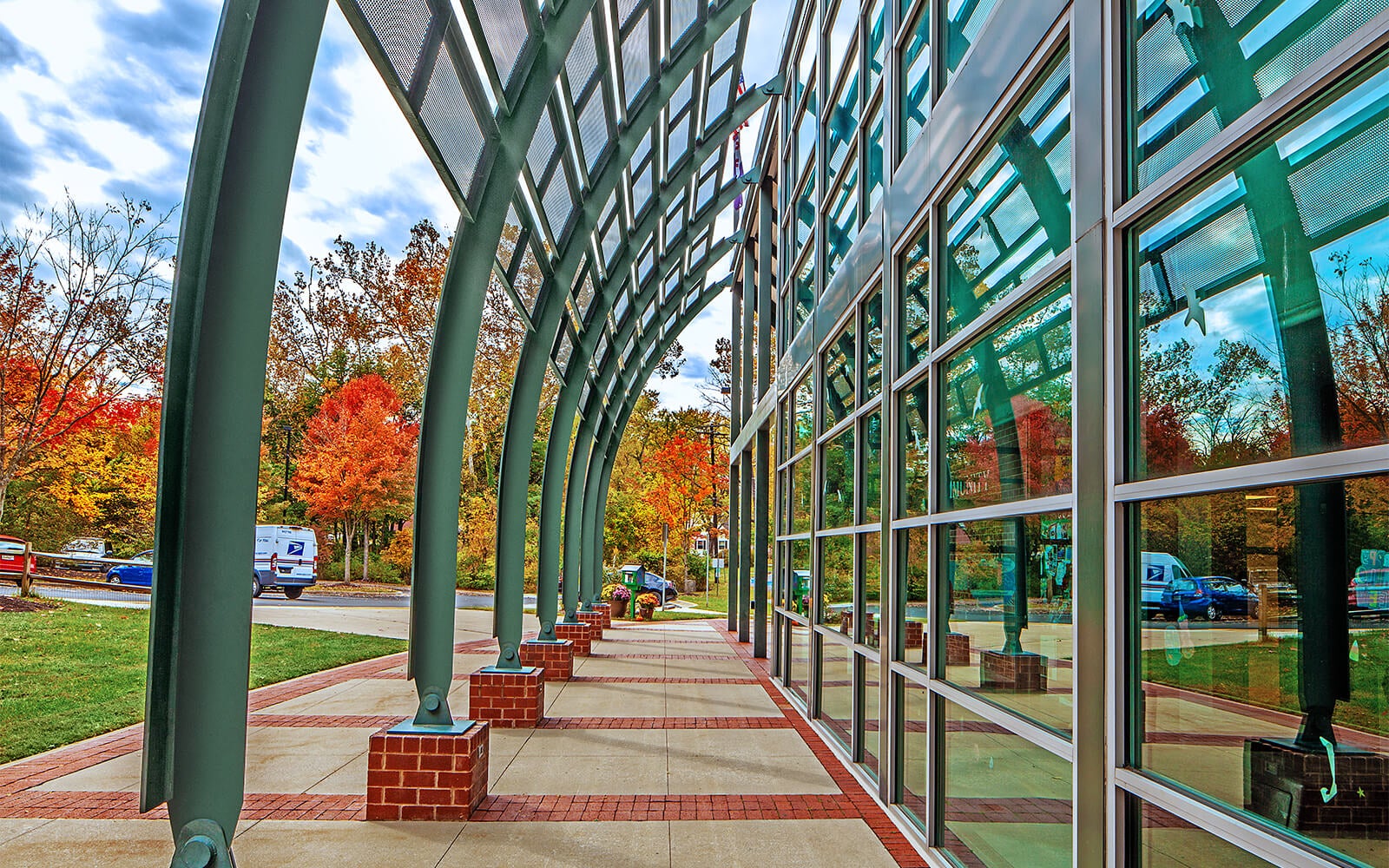Curved metal and glass canopy outside a building, reflecting trees with autumn foliage, cars, and a person sitting on a bench.