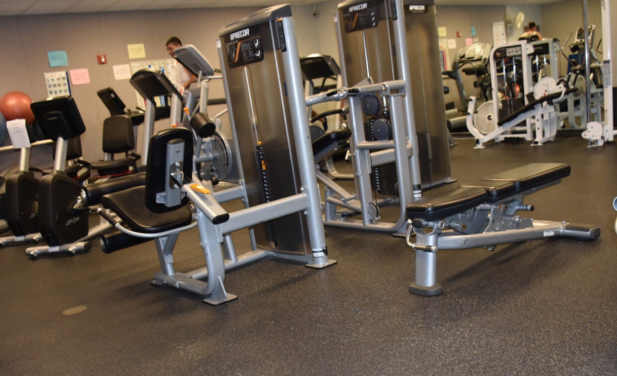 A gym with various exercise machines and equipment, including weight benches, seated row machines, and stability balls, arranged on a rubber floor.