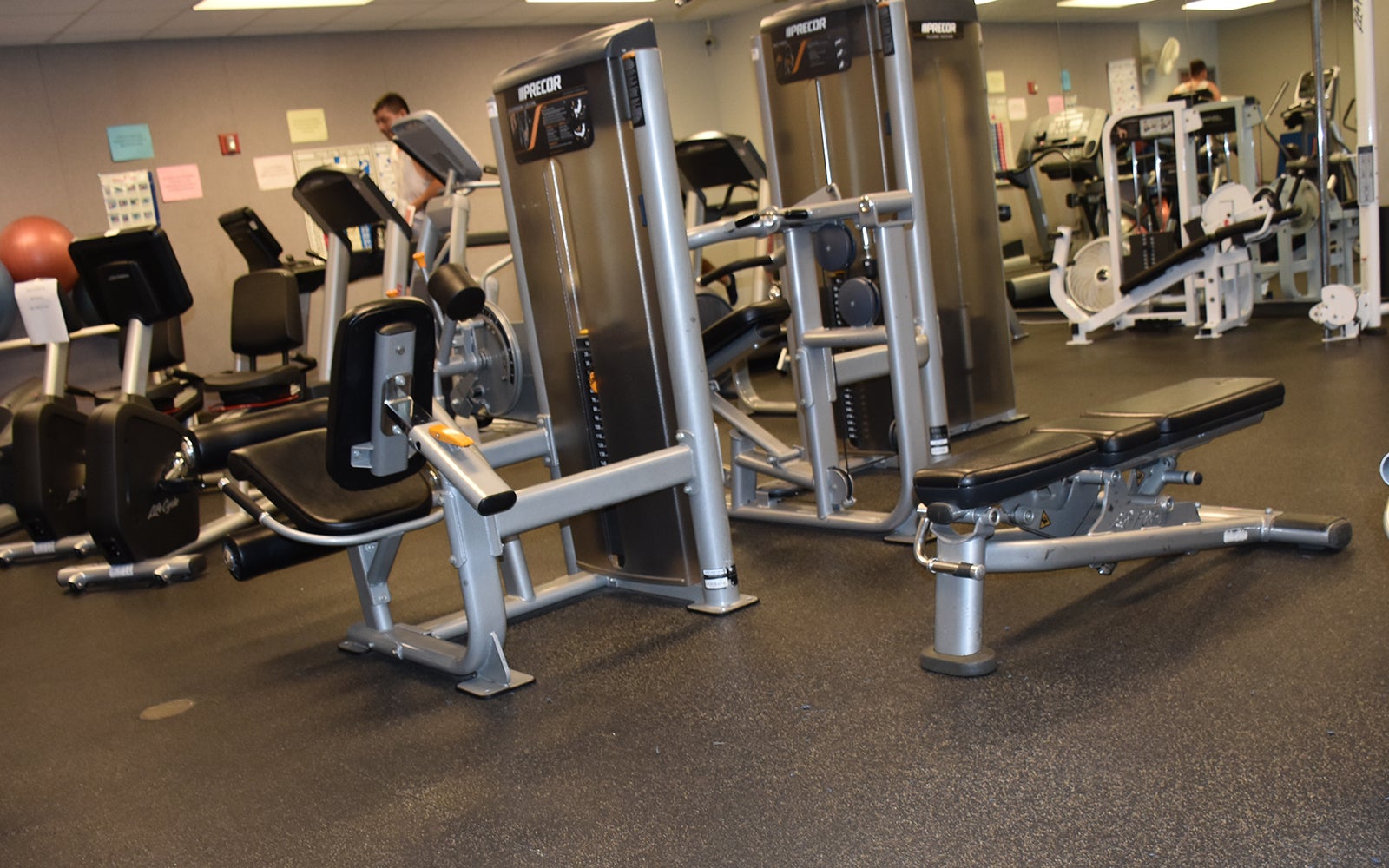 A gym with various exercise machines and equipment, including weight benches, seated row machines, and stability balls, arranged on a rubber floor.