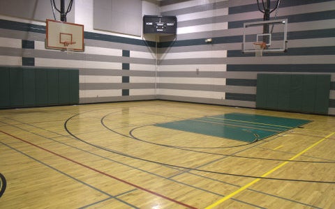 Empty indoor basketball court with wood flooring, striped walls, two basketball hoops, and a scoreboard in the corner.